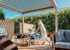 A woman setting an outdoor dining table under a luxury Instashade Technic 4m x 3m louvred pergola on a sunny timber deck.