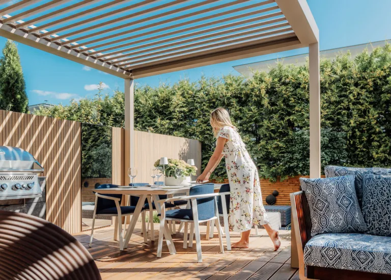 A woman setting an outdoor dining table under a luxury Instashade Technic 4m x 3m louvred pergola on a sunny timber deck.