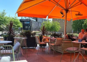A vibrant orange Instashade Nova centre post commercial parasol with integrated heaters providing shade on a modern urban restaurant terrace at the London Television Centre
